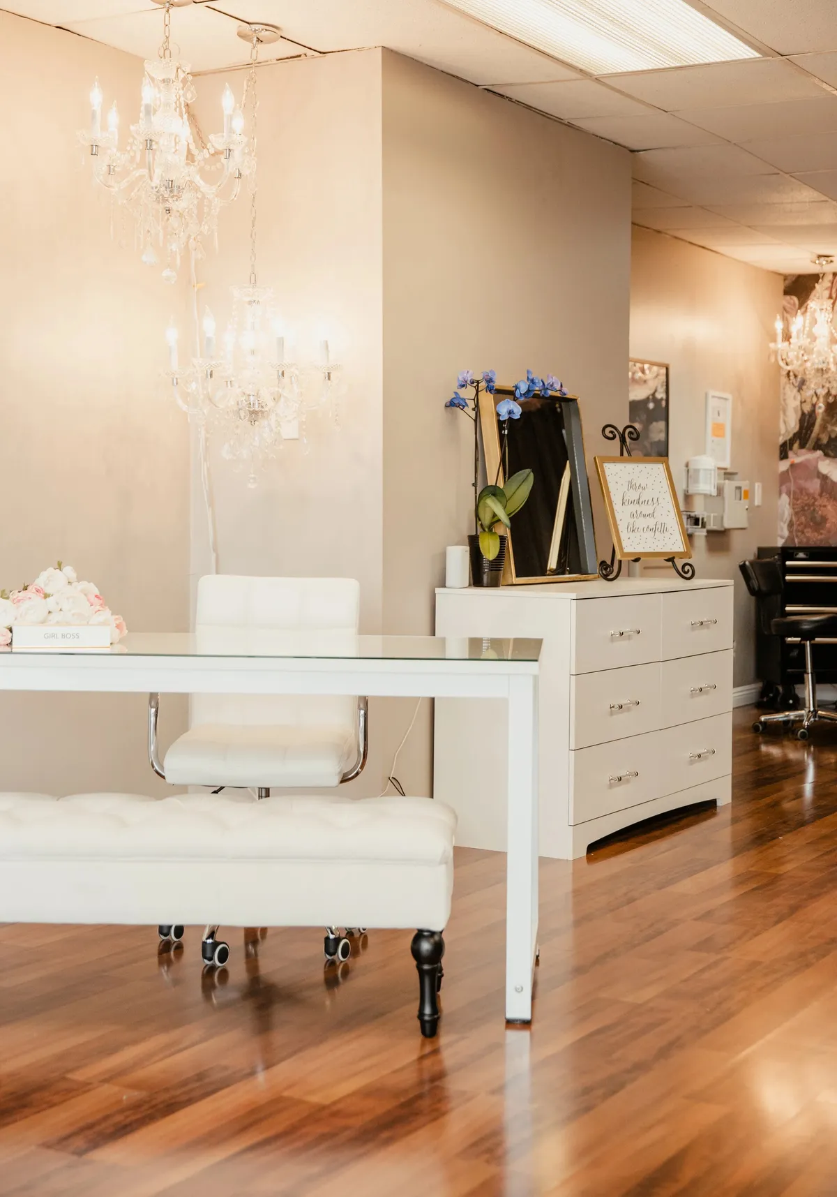 Reception desk at Open Circle Salon under two crystal chandeliers, with a white glass desk, tufted bench, and gold-framed mirror