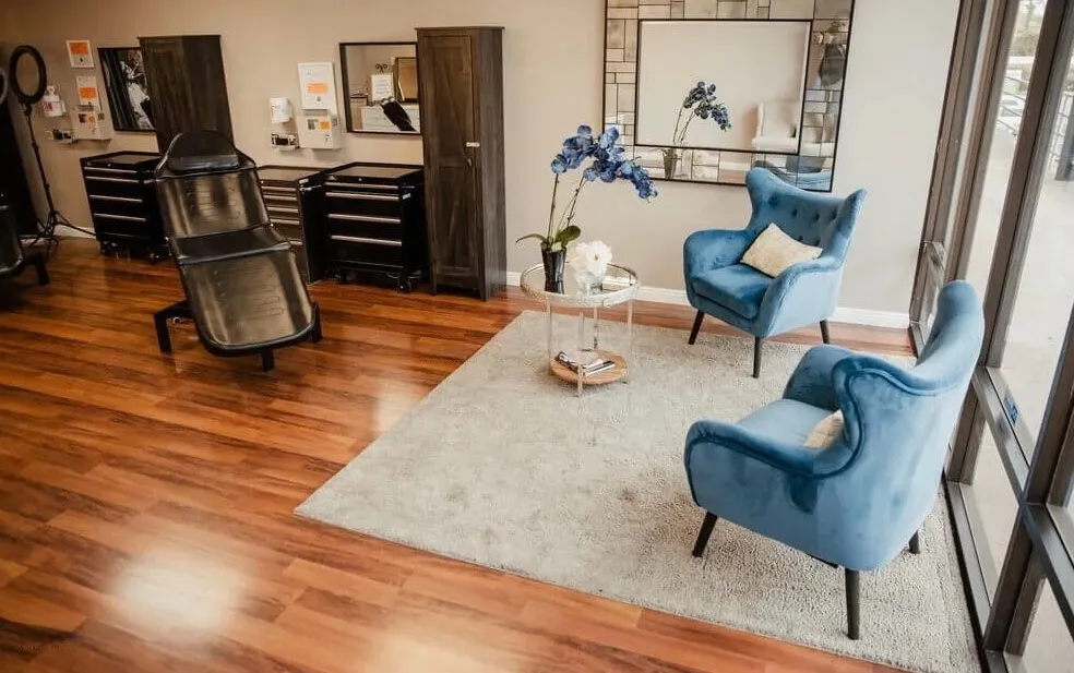 Seating area at Open Circle Salon with two teal velvet armchairs and a glass table on a cream rug, beside the treatment floor