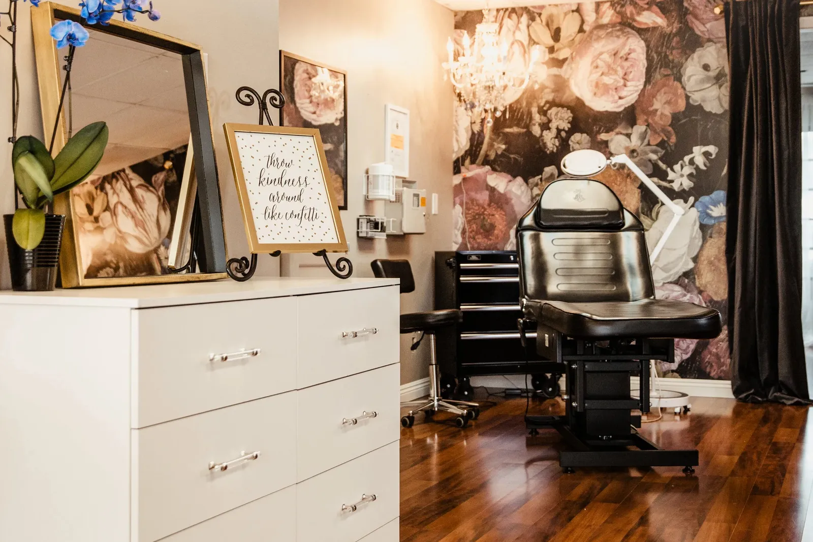 Treatment area at Open Circle Salon in Huntington Beach, with a reclining lash bed against a dark floral feature wall, a crystal chandelier overhead, and a gold-framed mirror on the dresser