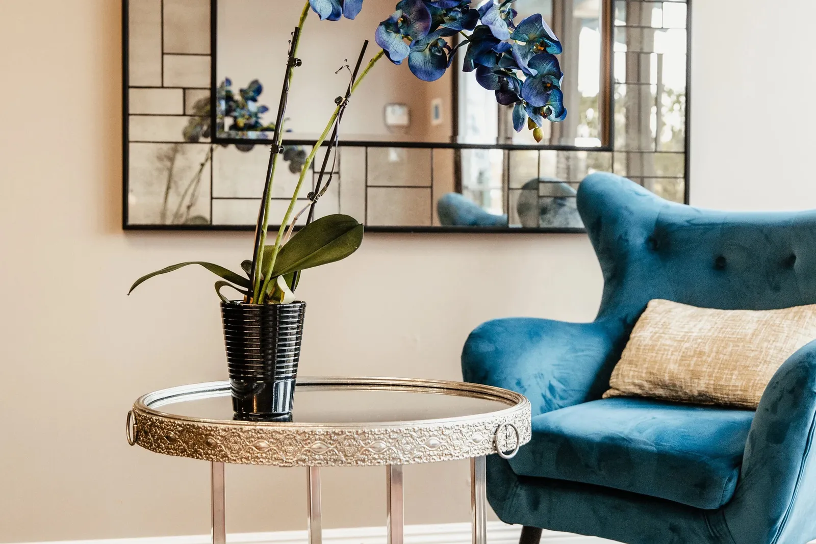 Waiting area at Open Circle Salon with a teal velvet armchair, a mirrored side table holding blue orchids, and brow reference books on the lower shelf
